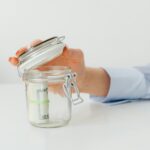 A hand places money in a glass jar on a white table, symbolizing savings.